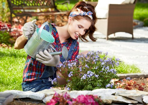 A young woman with light skin, brown hair tied with a colorful headband, wearing a red and blue plaid shirt and grey gardening gloves, is kneeling on the lawn in a garden during daytime. She is watering a flowering purple and white garden bed using a metal watering can. The garden area is bordered by flat stones, with a lush green lawn and various plants visible in the background, including a hedge, small trees, and a wooden garden bench. The outdoor space appears well-maintained, with natural sunlight illuminating the scene, and the garden showcases a mix of flowering plants and landscaped elements typical of residential gardens in Mayfair. The image reflects outdoor gardening activities, supporting professional gardening services focused on flower bed care and garden maintenance.